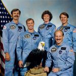 Five astronauts in blue jumpsuits pose with model American eagle in front of US flag