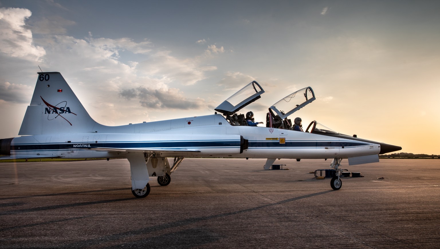 A NASA training jet, white with blue trim, is seen with its two cockpit windows open against the Florida sky at dusk.
