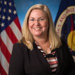 A white female with blond hair and is in a suit poses for a picture in front of the American flag and the NASA flag