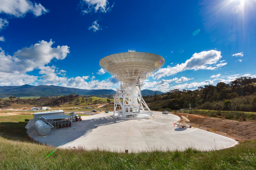 A 34-meter (111-foot) antenna points toward a bright blue sky. Mountains and several clouds can be seen in the background.