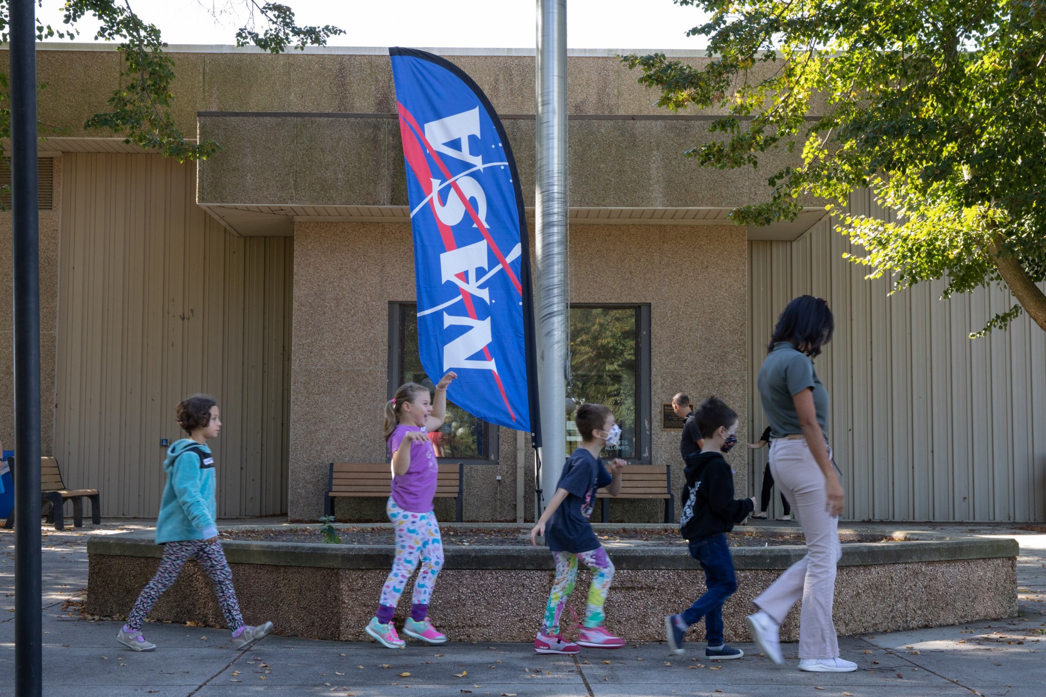 four children follow behind a woman in tan pants and a gray polo shirt outside a concrete building with a bright blue NASA feather flag in the background