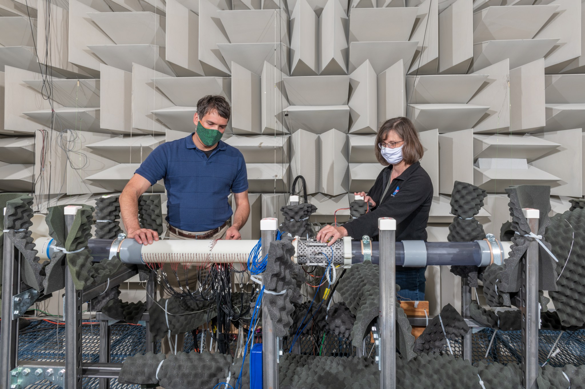 : A man and a woman wearing masks work on equipment inside a NASA acoustic facility. Large tan fiberglass wedges line the walls of the facility.
