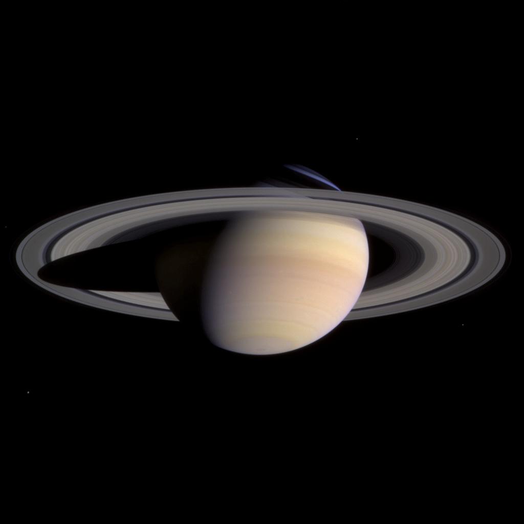 Saturn and its rings against the blackness of space. Saturn is a pale beige, with slightly varying stripes. At the top is a sliver of blue light.
