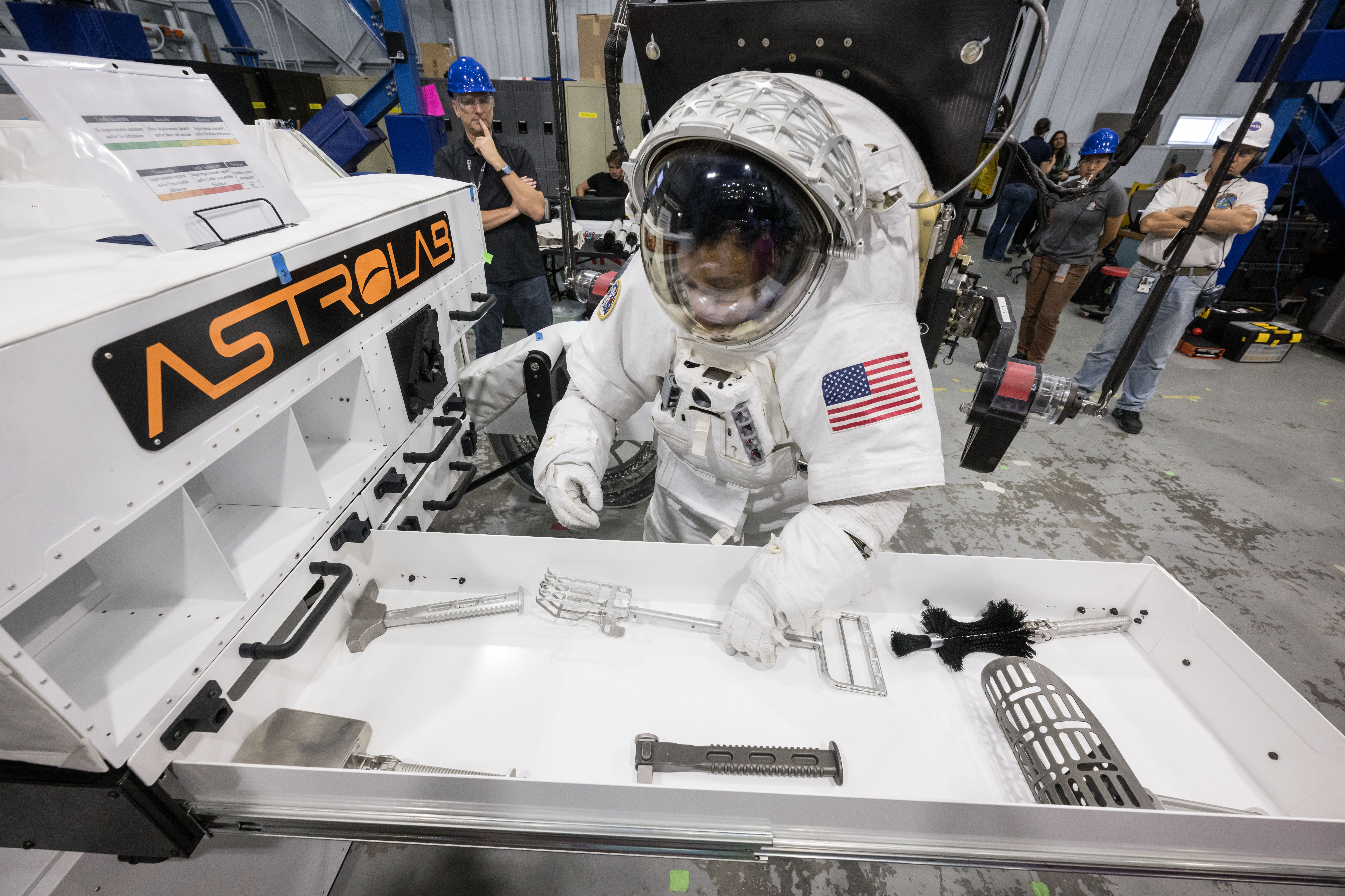 NASA astronaut Jessica Watkins is wearing a white spacesuit with the American flag on her left shoulder. She is pickup up a lunar geology tool from...