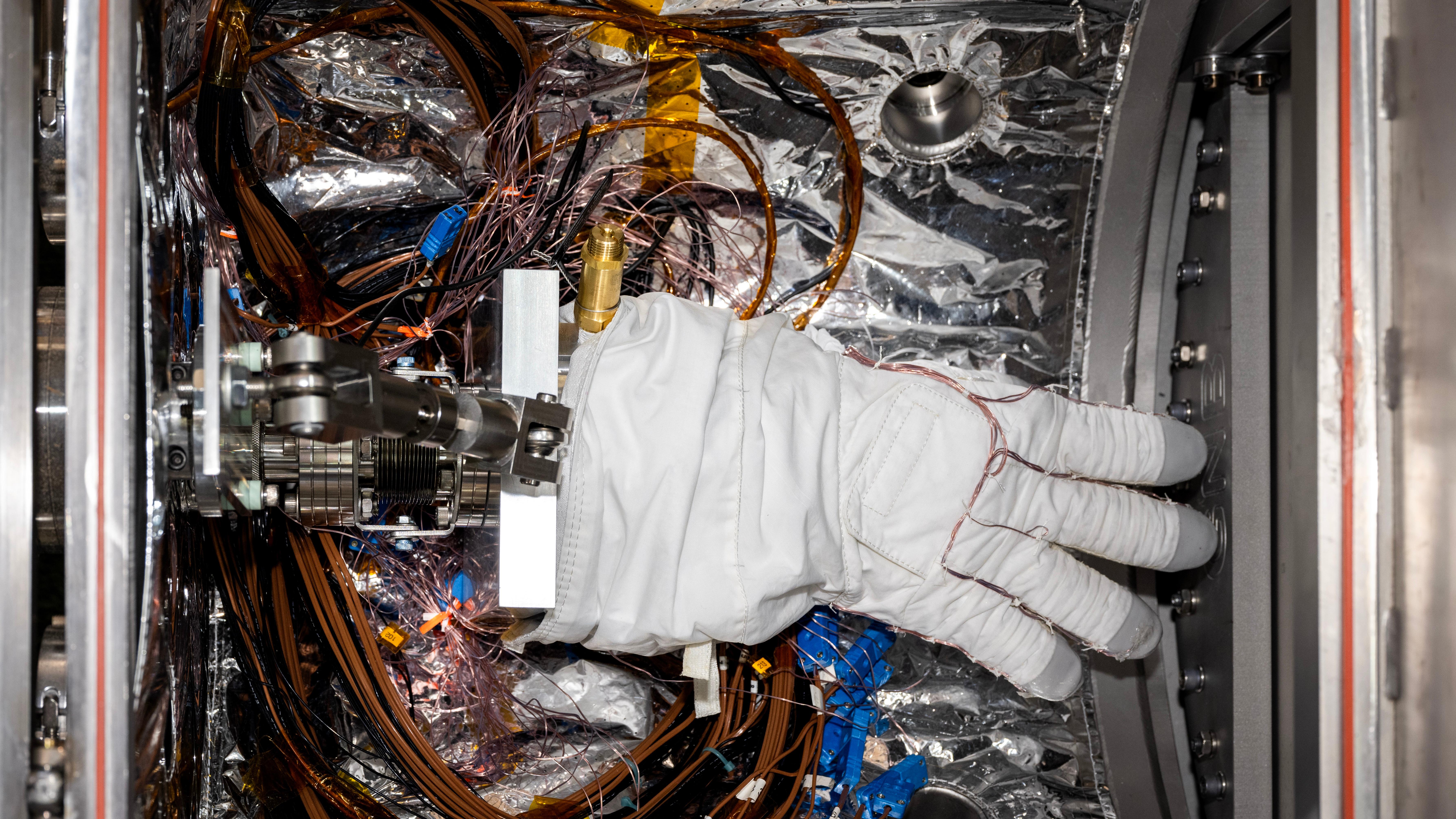 A bulky white spacesuit glove with gray fingertips sits inside a small chamber lined with silvery material. Multiple wires wrap around the fingers of the glove and loop around the floor of the chamber beneath the glove.