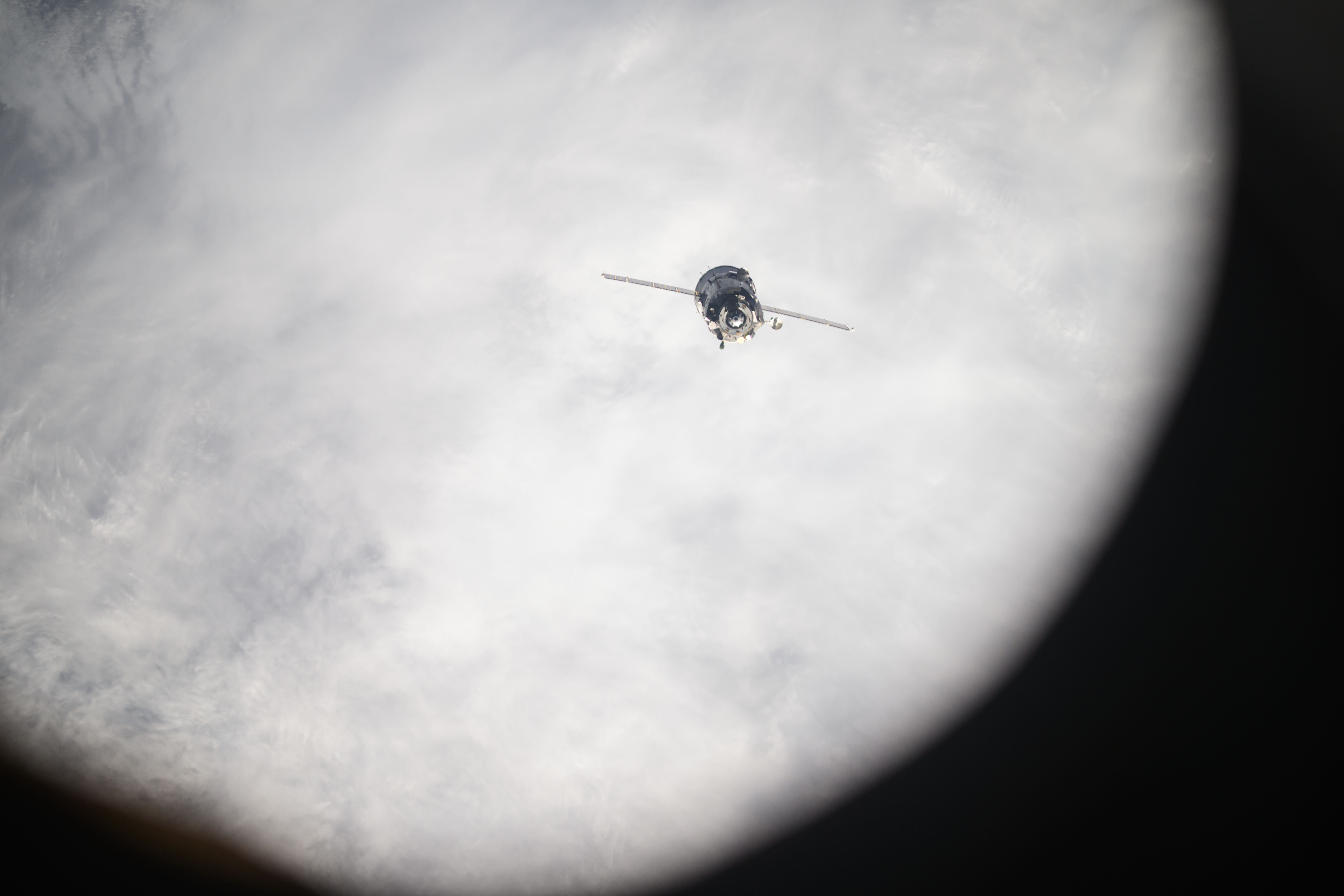 The Soyuz spacecraft looks like a cylinder with two lines stretching outward from it. It is oriented toward Earth, which is covered with white clouds.